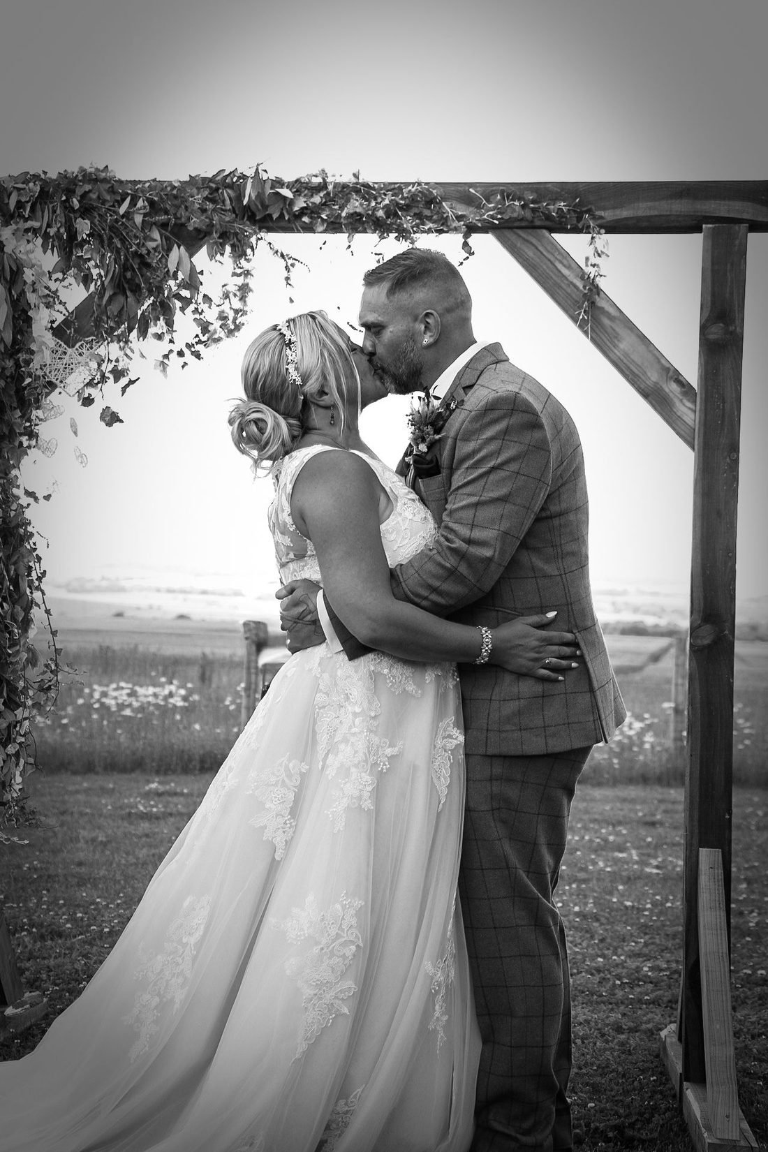 First kiss beneath a flower-covered wedding arch, in black and white