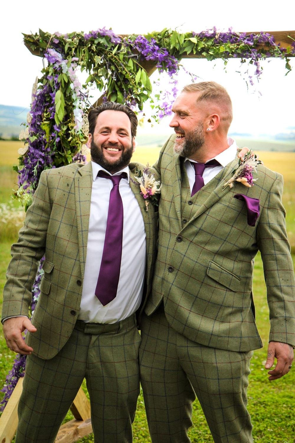 Groomsmen in green tweed laughing beneath the flower arch