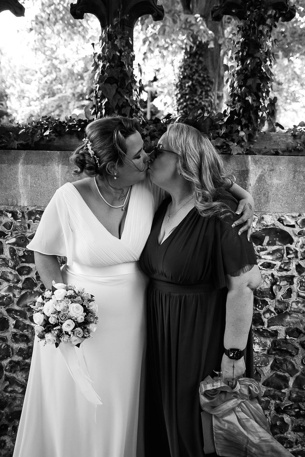 Two brides kissing beneath an ivy-covered lychgate, in black and white
